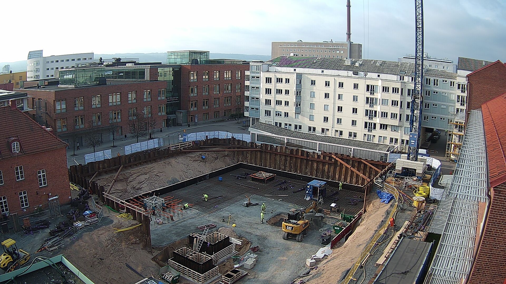 Byggarbetsplatsen sett från nuvarande Science Park House, med bland annat högskolan i bakgrunden. En gul grävmaskin och byggarbetare är på plats och jobbar.