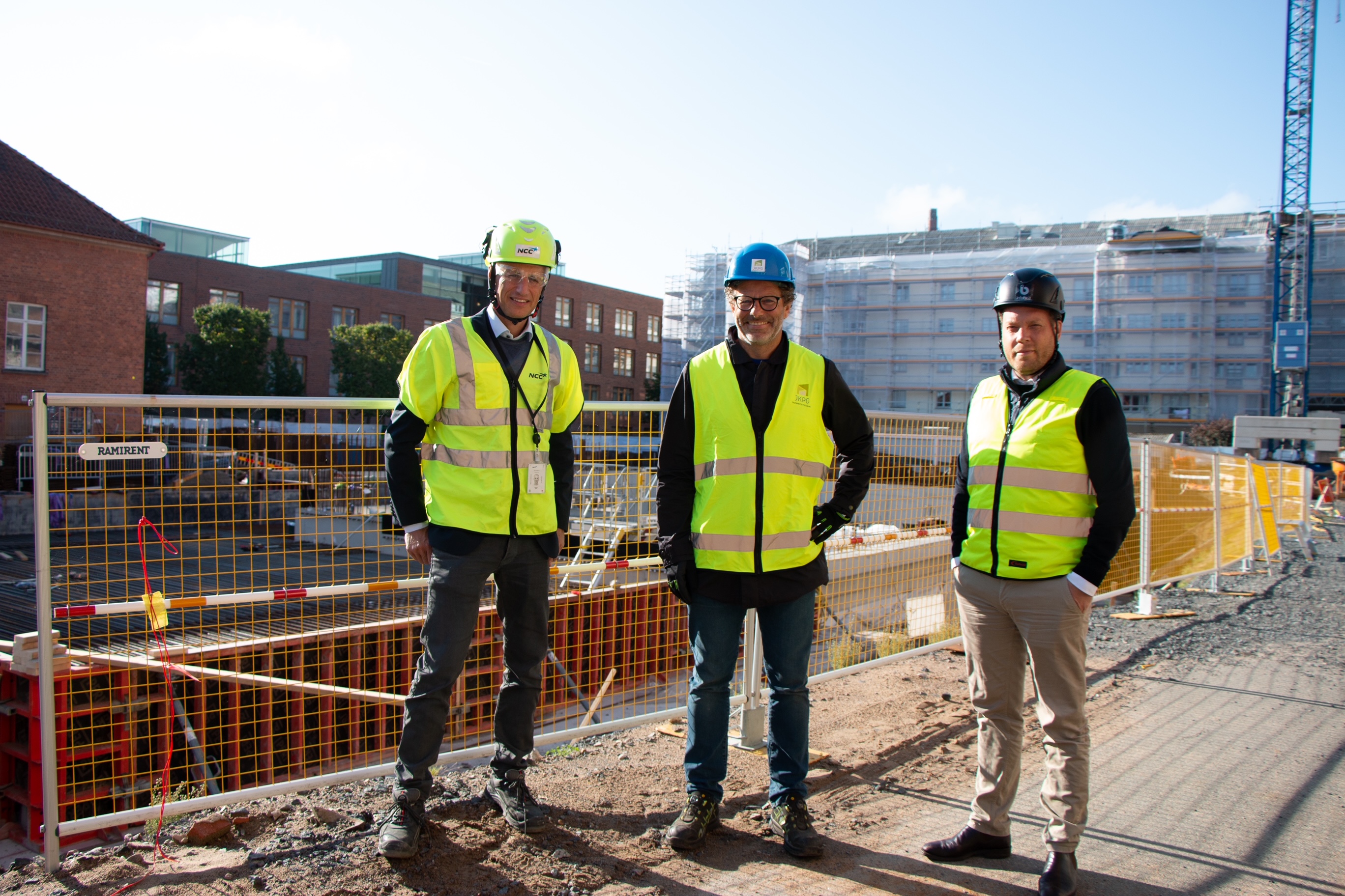 De 3 parterna står i solen framför byggarbetsplatsen.