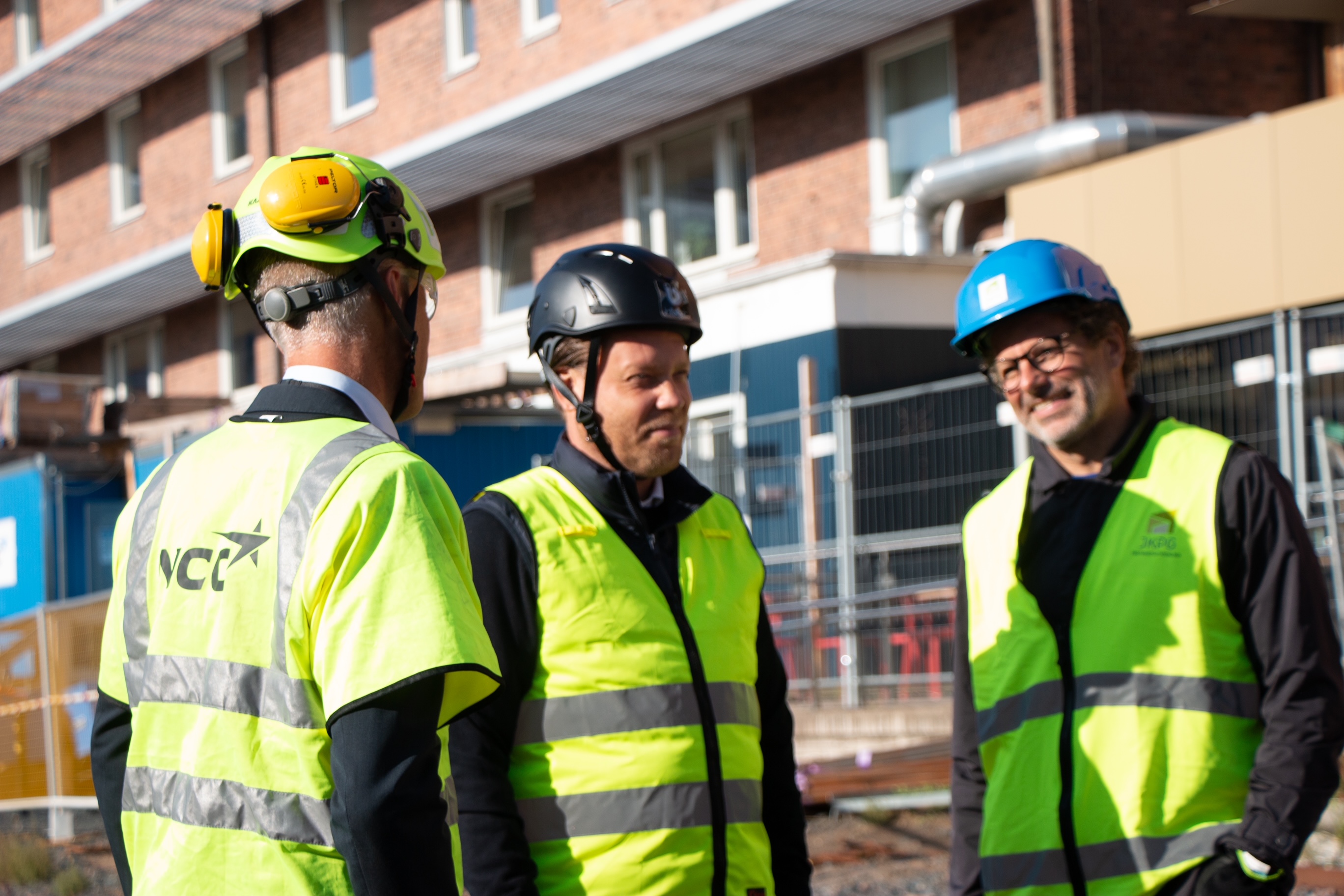 Anders Saltin, Magnus Olsson och Dan Friberg på byggarbetsplatsen. Parterna står vända mot varandra, pratar och skrattar i solen.