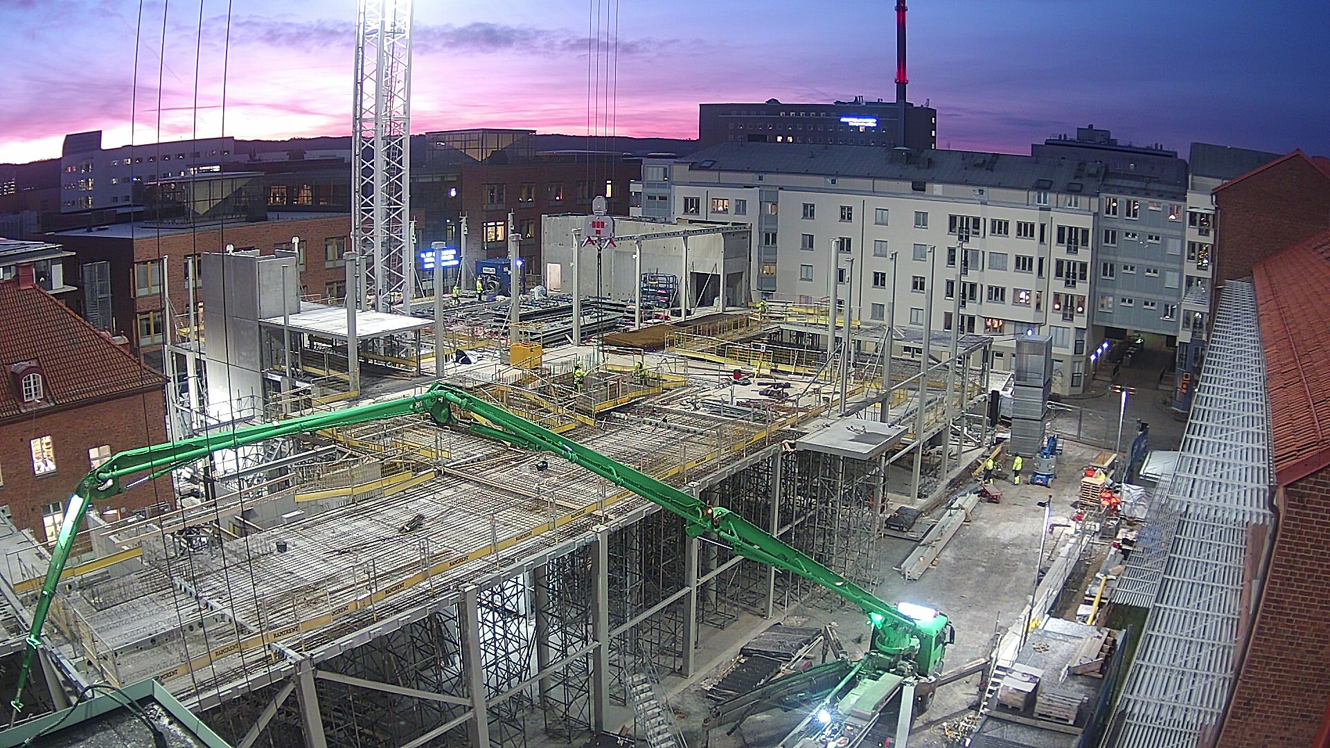 Byggarbetsplatsen 2021-12-21. Det är morgon och himlen är rosa från solen som är på väg upp. Byggarbetare är på plats och reser stommen.