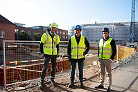 De 3 parterna står i solen framför byggarbetsplatsen.